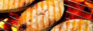 Three pieces of fish steak are being grilled over an open flame, showing visible grill marks on the surface.