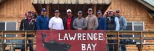 Ten men stand on a wooden lodge balcony behind a large sign that reads “Lawrence Bay Lodge” with a fish illustration.