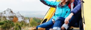 Two people sit at the entrance of a yellow tent on a hillside, looking and pointing at the landscape ahead.