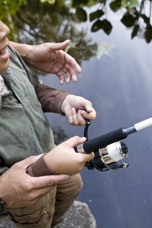 Adult helping a child hold and use a fishing rod near the edge of a body of water.