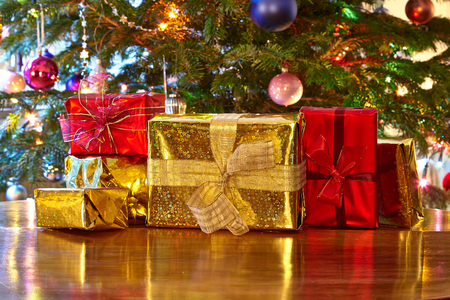 Several wrapped gifts in gold and red paper sit on a wooden table in front of a decorated Christmas tree.