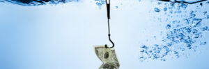 A fishhook holding a folded U.S. dollar bill is submerged in clear blue water with bubbles.