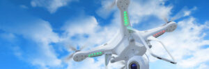 White quadcopter drone with camera flying against a blue sky with scattered clouds.