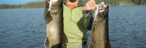 Man in sunglasses and a hat holds two large fish while standing on a boat in a lake with forest in the background.