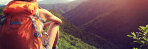 Person with an orange backpack sitting on a rock, overlooking a green mountain landscape under a bright sky.