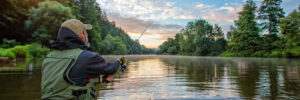 Person fishing in a river at sunrise, surrounded by trees and calm water under a partly cloudy sky.
