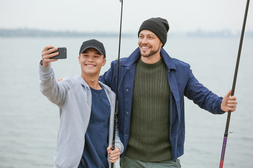 Two men fishing, each holding fishing rods. One is taking a selfie of them both, representing that the two are enjoying their fishing trip