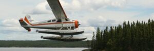A floatplane flies low over a lake near a forested shoreline under a partly cloudy sky.