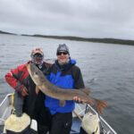 Two people on a boat hold a large fish, smiling, with cloudy skies and open water in the background.