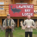 Two men hold large moose antlers in front of a sign for Lawrence Bay Lodge, standing by a wooden building.