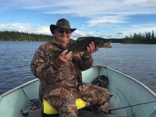 Man in camouflage outfit holding a fish on a boat in a lake, wearing a brown hat and smiling. Forested shoreline in the background.
