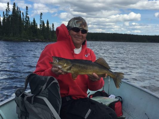 Person in a red hoodie and camouflage cap holds a large fish in a boat on a lake, surrounded by trees and a cloudy sky.