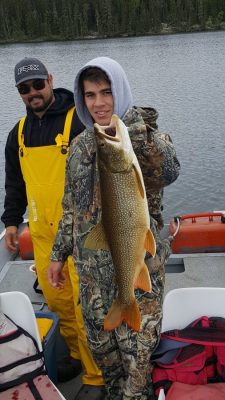 Two people on a boat with a large fish. One wears camo, holding the fish. The other in yellow rain gear stands nearby. Lake and trees behind.