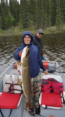 Two people in a boat on a lake; one holds a large fish, both smiling. Forested shoreline in the background.