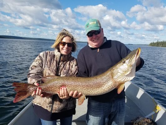 Two people on a boat holding a large fish, with a lake and cloudy sky in the background.