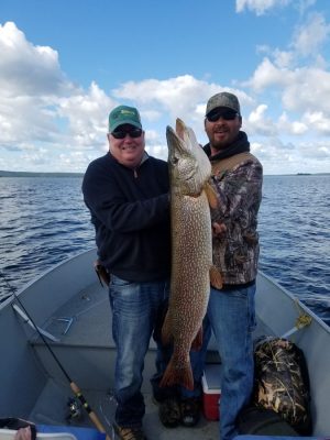 Two men on a boat holding a large fish, smiling at the camera. The background shows a lake under a partly cloudy sky.