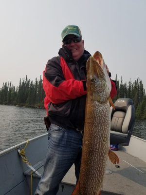 Person holding a large fish on a boat, wearing a red and black jacket and a green cap, with trees and water in the background.