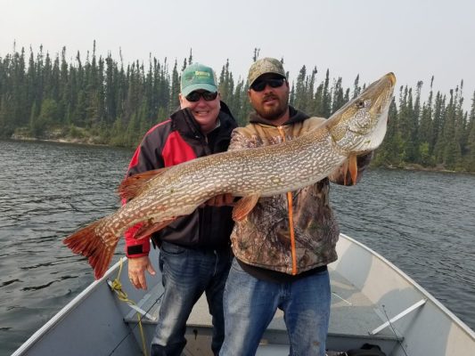 Two people on a boat holding a large fish, with a backdrop of water and trees.