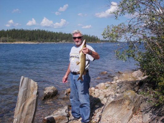 Man holding a fish by a lake, wearing jeans and a T-shirt, surrounded by rocks and trees under a partly cloudy sky.