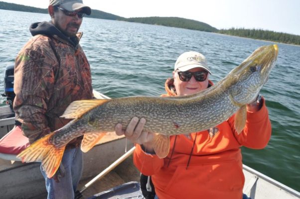 Person in an orange hoodie holding a large fish on a boat, with another person in a camo jacket nearby. Lake and trees in background.