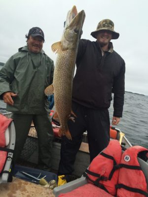 Two people on a boat holding a large fish. One wears a green rain suit, the other a dark jacket and hat. Life jackets are visible.
