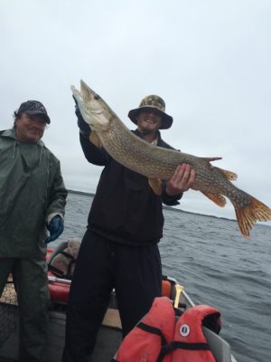 Two people on a boat; one holds a large fish horizontally, while the other stands nearby wearing a rain jacket.