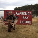 Person kneeling behind a moose head in front of a "Lawrence Bay Lodge" sign with a lake and trees in the background.