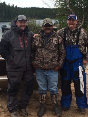 Three men in outdoor gear stand smiling on a sandy lakeshore with trees and boats in the background.