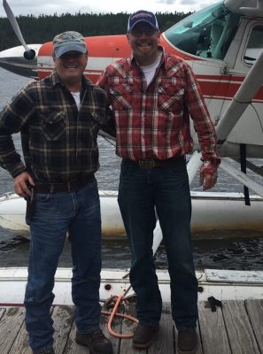 Two people in plaid shirts and caps stand on a dock beside a floatplane on the water.