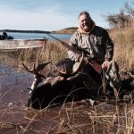Person in camouflage holding a rifle, sitting on a moose by a lakeside with a boat in the background.