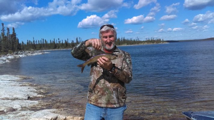 Man in camouflage jacket holding a fish by a lakeshore, with blue sky and clouds in the background.