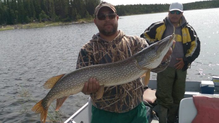 Man in camouflage jacket holding a large fish on a boat with another person standing behind him on a lake.