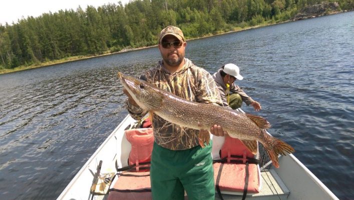 Man standing in a boat on a lake holding a large fish, with another person seated and trees in the background.