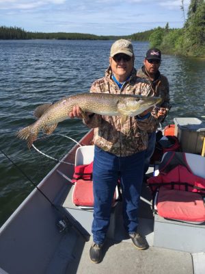 Person holding a large fish on a boat, with another person in the background. Lake and trees visible.
