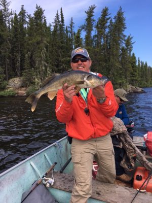 Person in a boat holding a large fish, wearing sunglasses and an orange jacket, with forested shoreline in the background.