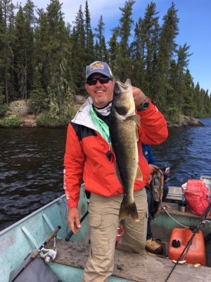 Person in a red jacket and cap holding a large fish on a boat, surrounded by water and tall evergreen trees in the background.