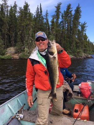 Person in a red jacket and cap holding a large fish on a boat with a wooded shoreline in the background.