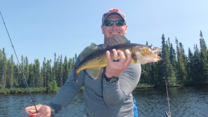 Person holding a fish while standing on a boat near a forested lake, wearing a cap and sunglasses, smiling at the camera.
