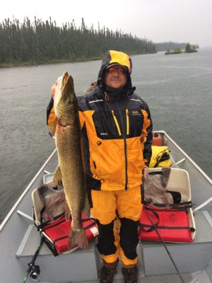 Person in yellow and black rain gear holding a large fish on a boat in a rainy lake setting.