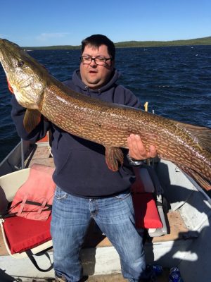 Person holding a large fish on a boat, with a lake and blue sky in the background.