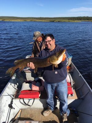 Two people on a boat; one is holding a large fish, while the other is in the background near the boat controls on a lake.