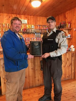 Two men standing in a wooden store. One holds a plaque while both are smiling. Fishing lures are displayed on the wall behind.