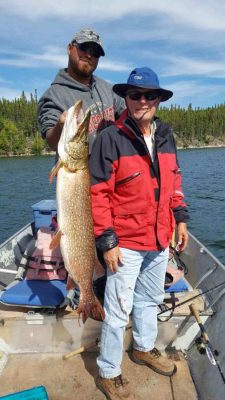 Two men on a boat holding a large fish, with a backdrop of a lake and forest.