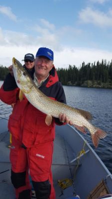 Man in red jacket holding a large fish on a boat with another person in the background; lake and trees visible.