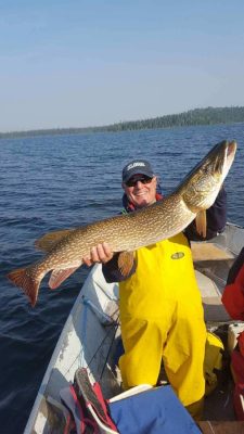 Person in a boat holding a large fish while wearing a yellow suit and cap on a lake with a forested shoreline in the background.