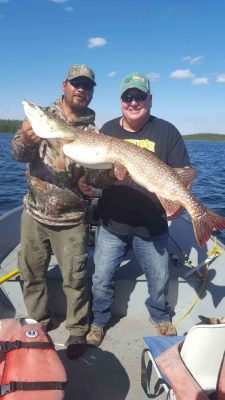 Two people on a boat holding a large fish, with a lake and blue sky in the background.