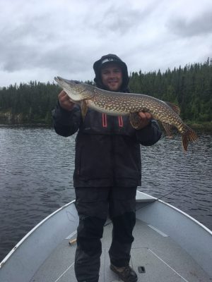 Person standing on a boat in a lake holds a large fish with both hands, surrounded by trees under a cloudy sky.
