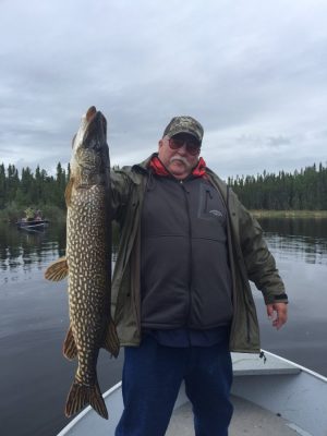 A person holding a large fish standing on a boat in a lake, surrounded by water and trees under a cloudy sky.