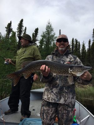 Two people on a boat, one holding a large fish, surrounded by trees under a cloudy sky.
