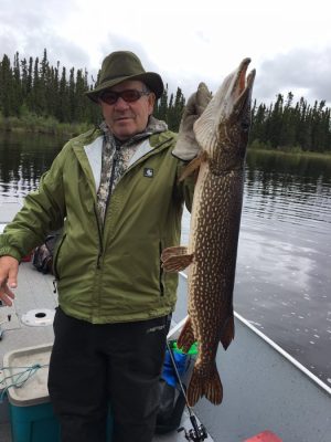 Person in green jacket and hat holding large fish on a boat, surrounded by water and trees in the background.